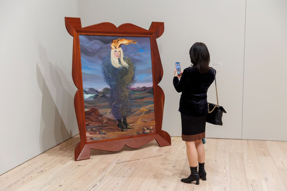 A person takes a photo of a painting, displayed in a large ornate frame at an art gallery.
