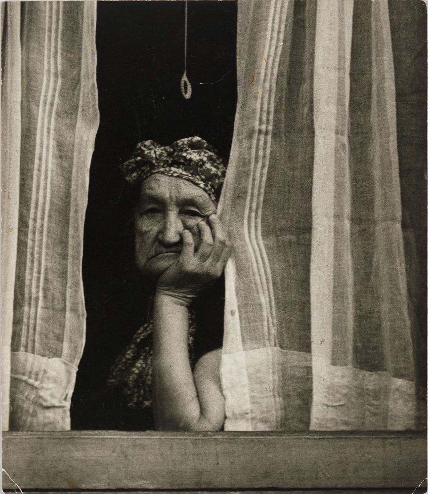 An elderly woman with a headscarf rests her chin on her hand, gazing out from behind striped curtains.