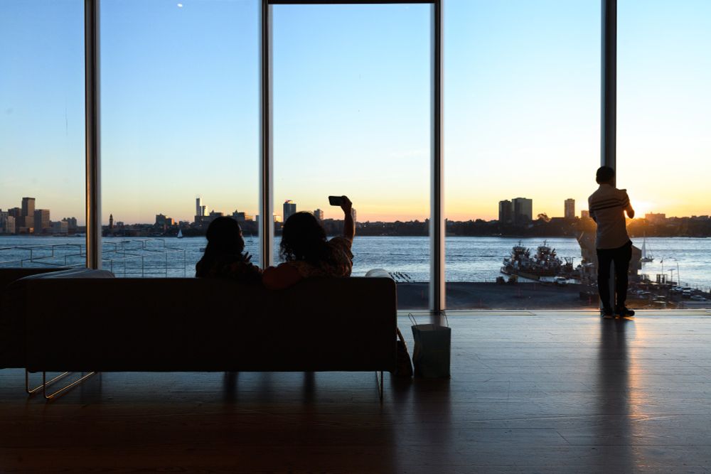 Two people sitting on a couch inside a room, silhouetted against a large window overlooking a river with a city skyline at sunset.