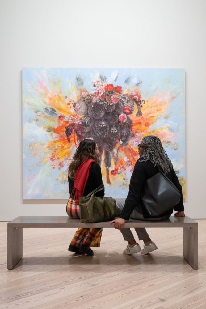 Two individuals sitting on a bench, viewing a large, colorful abstract painting in an art gallery.