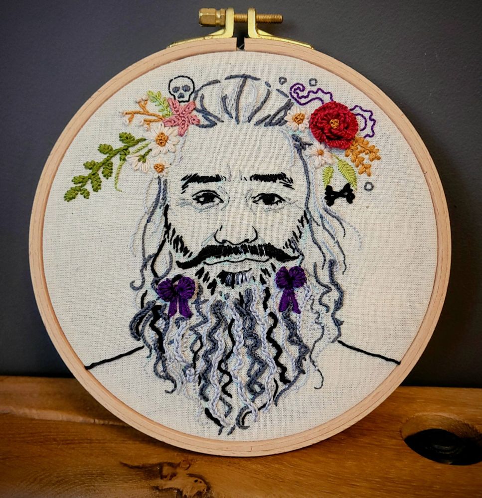 Embroidery in a hoop of a close-up of season 1 Edward smiling at the viewer with purple bows in his beard and an assortment of flowers in his hair)