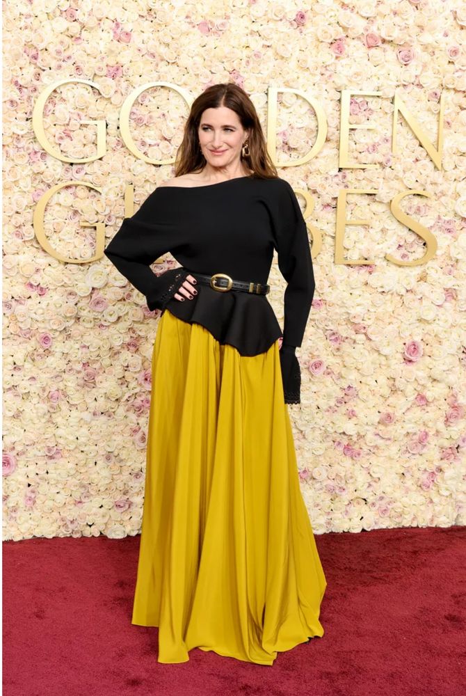 Woman posing on the Golden Globes red carpet in a golden yellow flowing skirt and an off the shoulder black long sleeved top with a belt. 