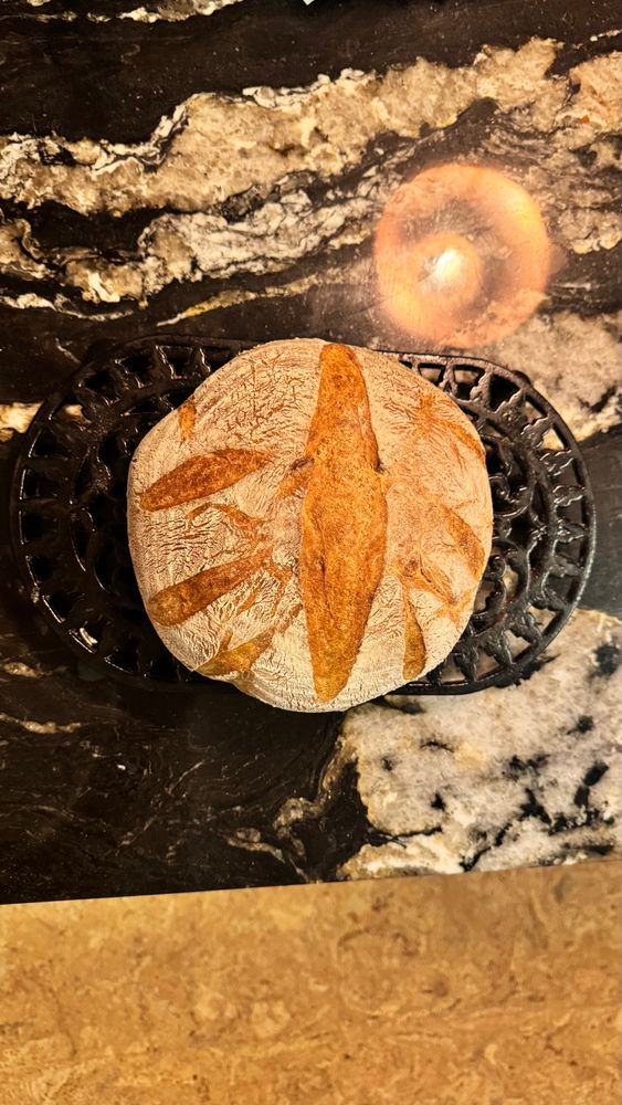 A golden brown round loaf of sourdough, cooling on a black iron trivet, sitting on a black and white marble counter top. The reflection of an overhead lamp can be seen in the marble of the counter top. 