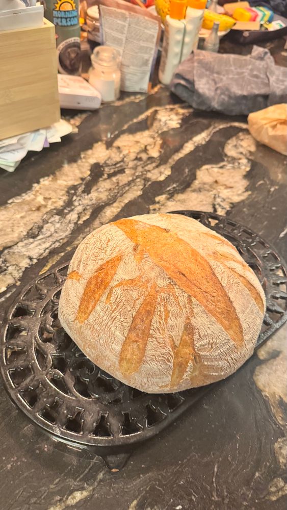A golden brown round loaf of sourdough, cooling on a black iron trivet, sitting on a black and white marble counter top. The slots cut to allow expansion stand out from the rest of the flour covered crust.