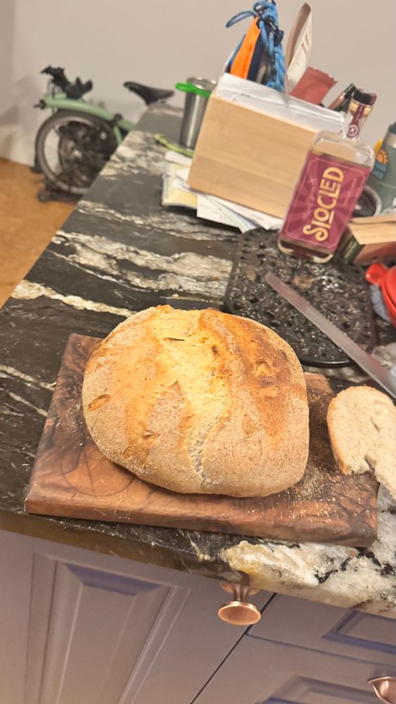 A golden round loaf of bread, on a dark walnut chopping board. A slice has been cut off the end. The bread knife is in the background, on the black Marble top. Blue kitchen cabinets underneath. 