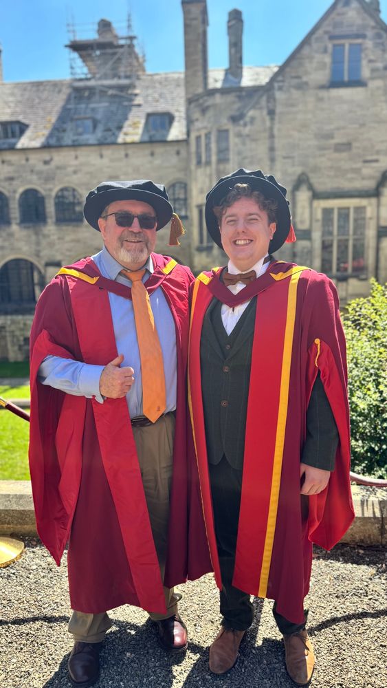 Two men in red and gold graduation gowns in a university courtyard. Professor Mullins on the right, giving his graduating PhD student Joseph Smith a hug in the inner quad at Bangor University.