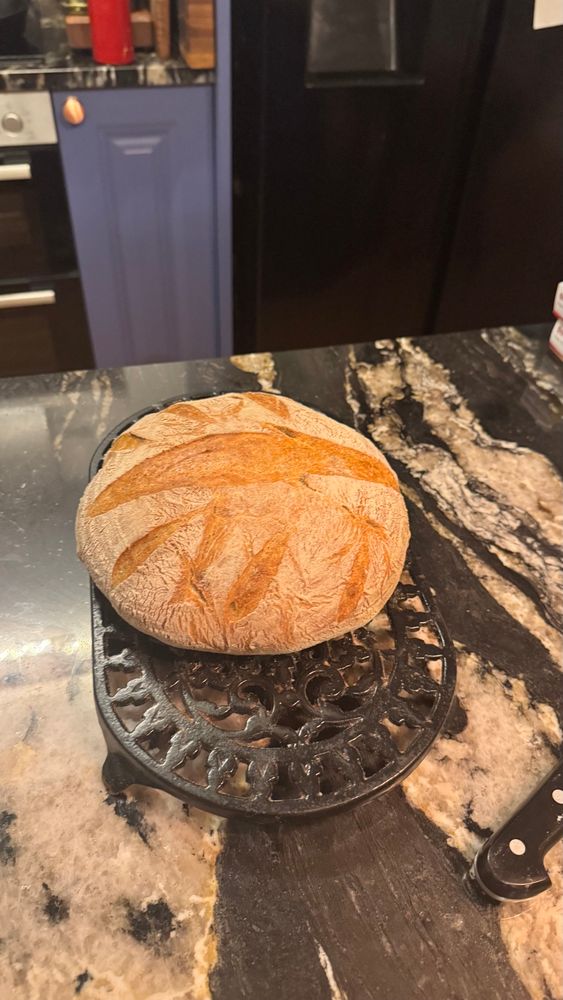 A golden brown round loaf of sourdough, cooling on a black iron trivet, sitting on a black and white marble counter top. 