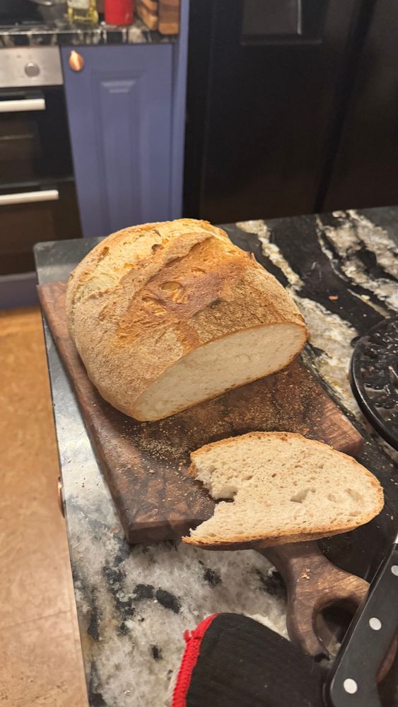 A golden round loaf of bread, on a dark walnut chopping board. A slice has been cut off the end. This view is from the front of the sliced end, showing the structure of the bread. All on the black and white Marble top of our kitchen island.