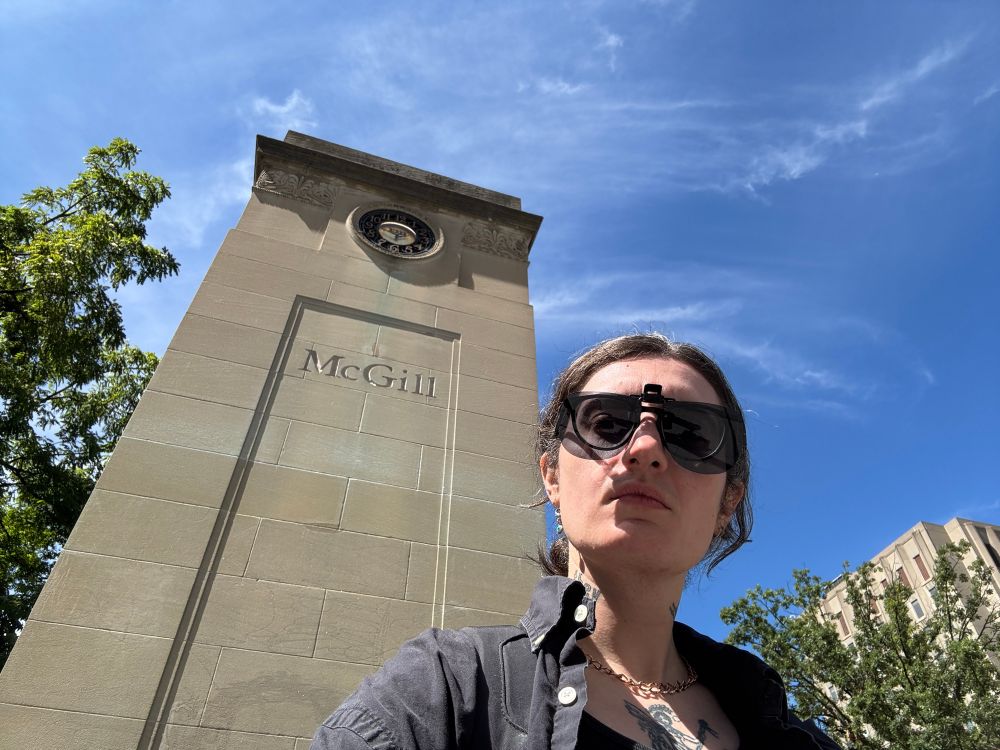 Sid on a sunny day standing in front of a structure that says McGill on it. it’s taken from a low angle and Sid is wearing an open button-down shirt and clip-on sunglasses so slightly giving dad on vacation energy, but it’s actually back to school 