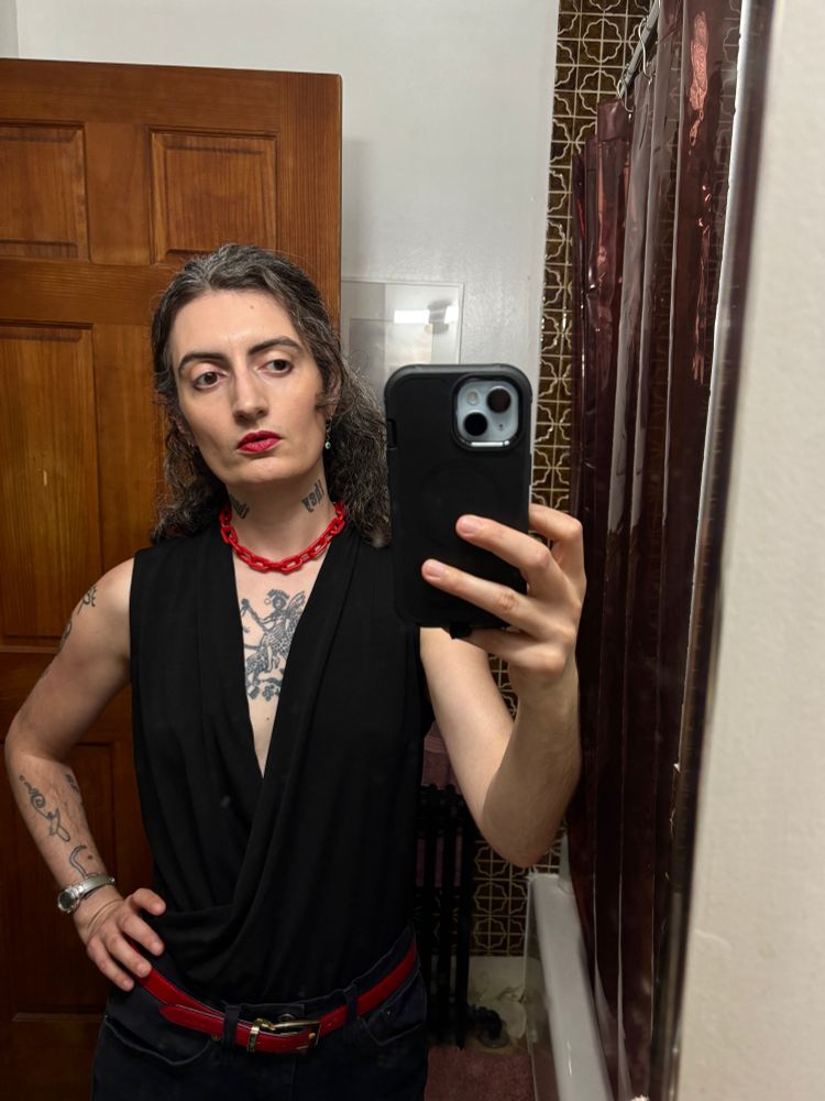 mirror selfie of sid wearing a black sleeveless top with a low neckline that shows their chest tattoo, black jeans, a red leather belt and a red plastic chain choker. they have actually done their makeup and hair. 