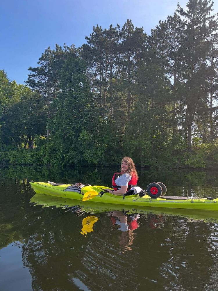 Dani, a middle aged woman, is sitting in a yellow kayak on the river and smiling back toward the camera. She’s wearing a red PFD and a white T-shirt. 