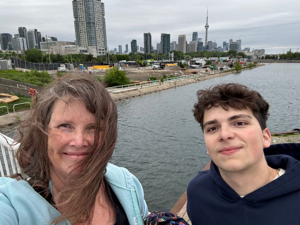 Dani, a smiling middle-aged mom with brown hair and dimples, and Simon, her 21 year old son with dark hair and eyes, in front of the Toronto waterfront skyline at Ontario Place, with the CN Tower and Lake Ontario in the background 