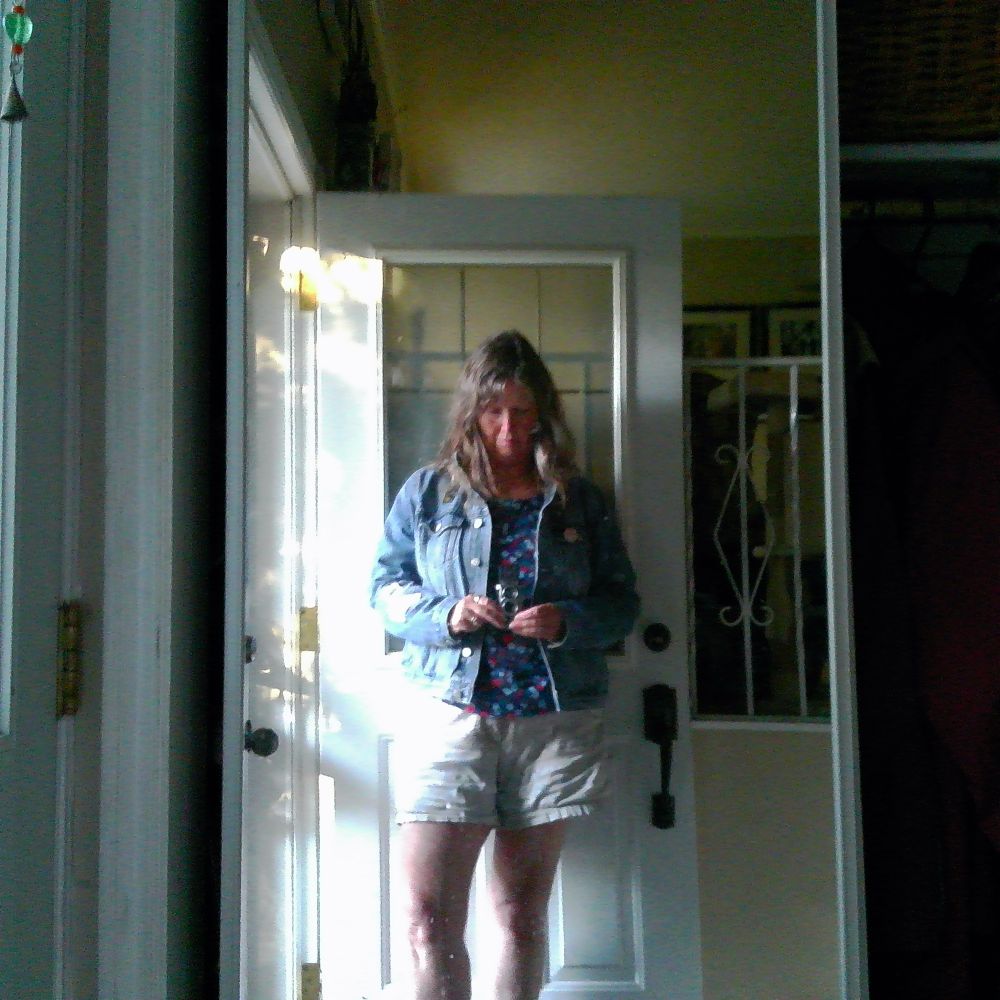 A mirror selfie of me, Dani, from a wide angle showing almost full body, wearing shorts and a jean jacket in front of an open door and looking down at the Chuzhao camera at belly button height. 