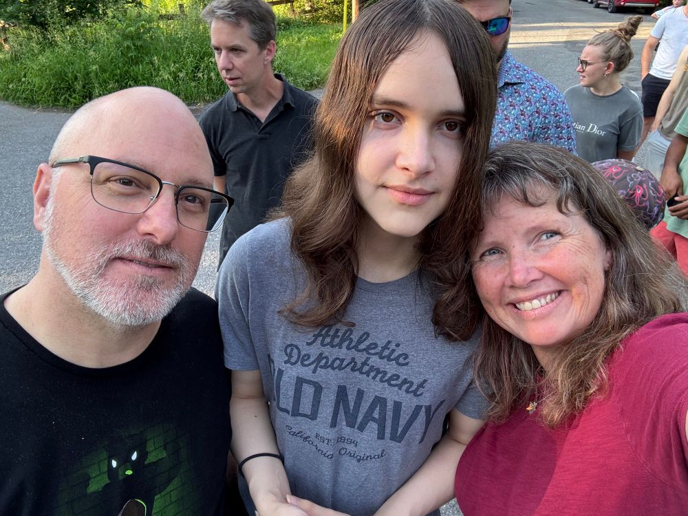 A family selfie with Dani, a smiling woman with freckles and dimples, her husband who has a salt and pepper beard and wears glasses and Lucas, an older teen with long hair. 