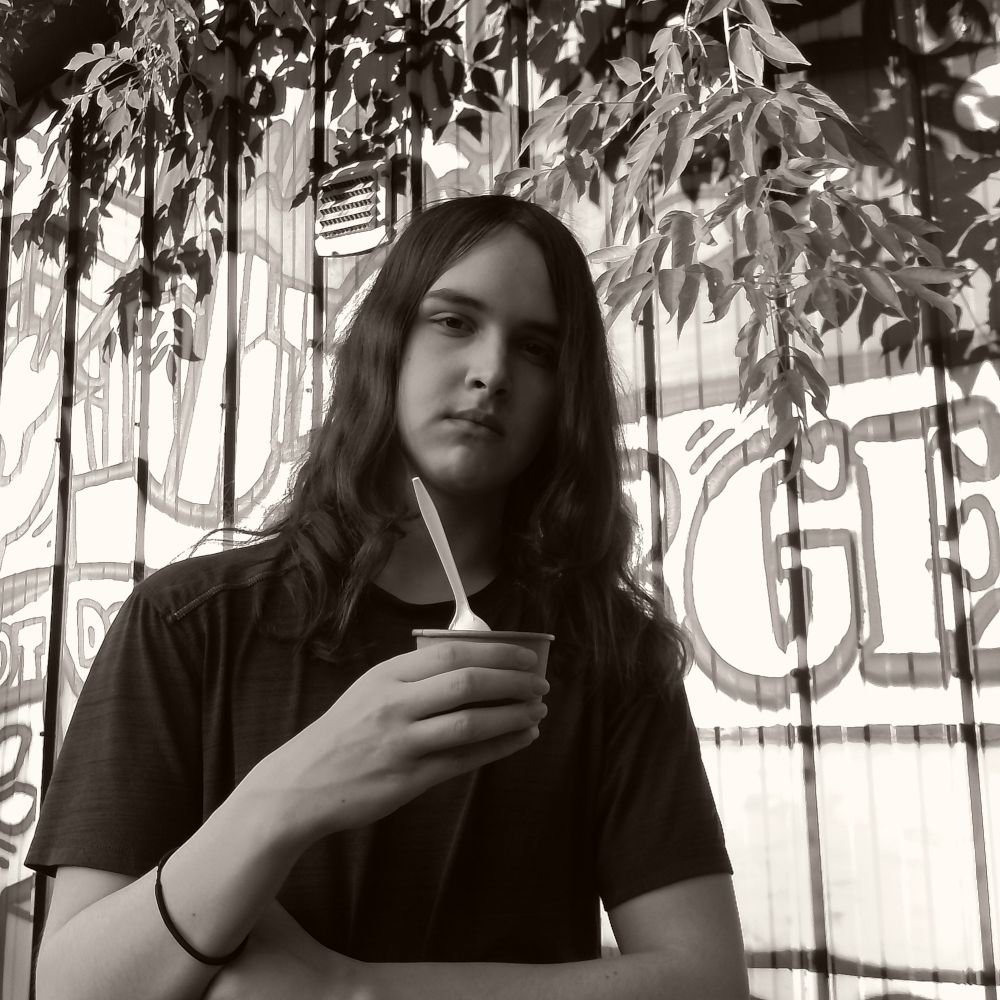 A black and white photo of Lucas, a 17 year old with long hair who might appear to be scowling but is holding an ice cream cup so can’t actually be all that grumpy. 