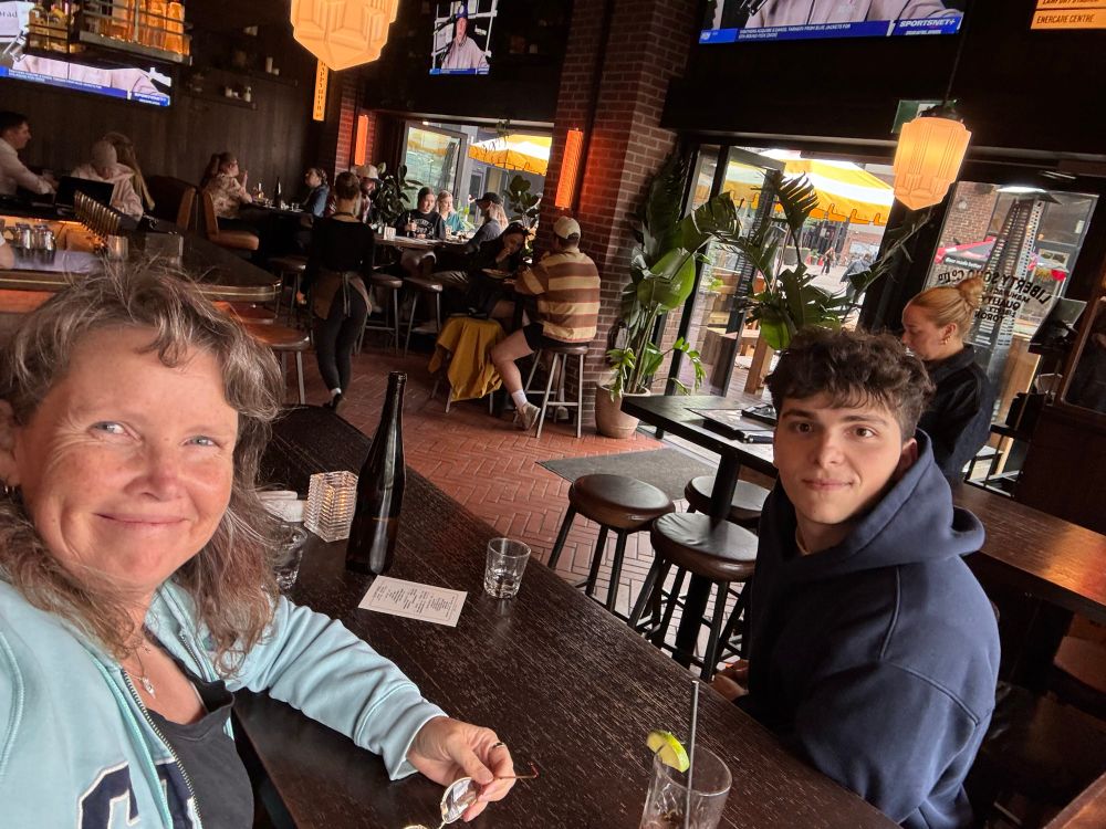 Dani, a smiling middle-aged mom with brown hair and dimples, and Simon, her 21 year old son with dark hair and eyes, at a table in the LibertySoho restaurant in the Liberty Village neighborhood of Toronto
