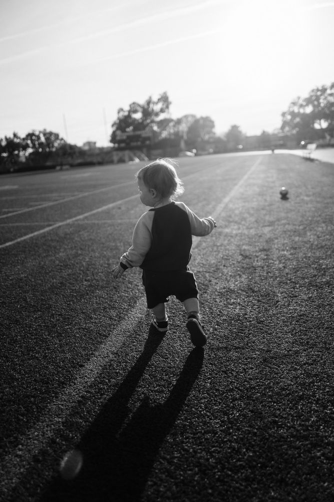 A black and white image of a toddler running on a high school football field, his back to camera. 