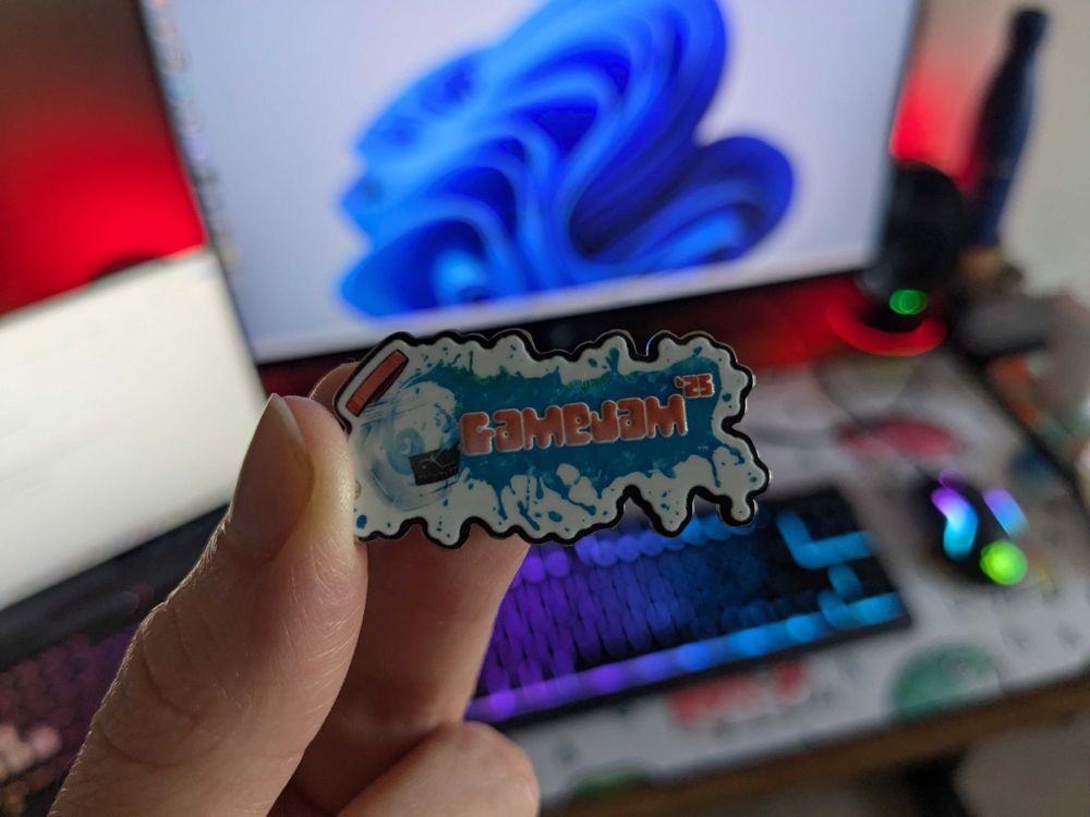 Someone holding a Game Jam 2025 pin badge in front of their desk.
