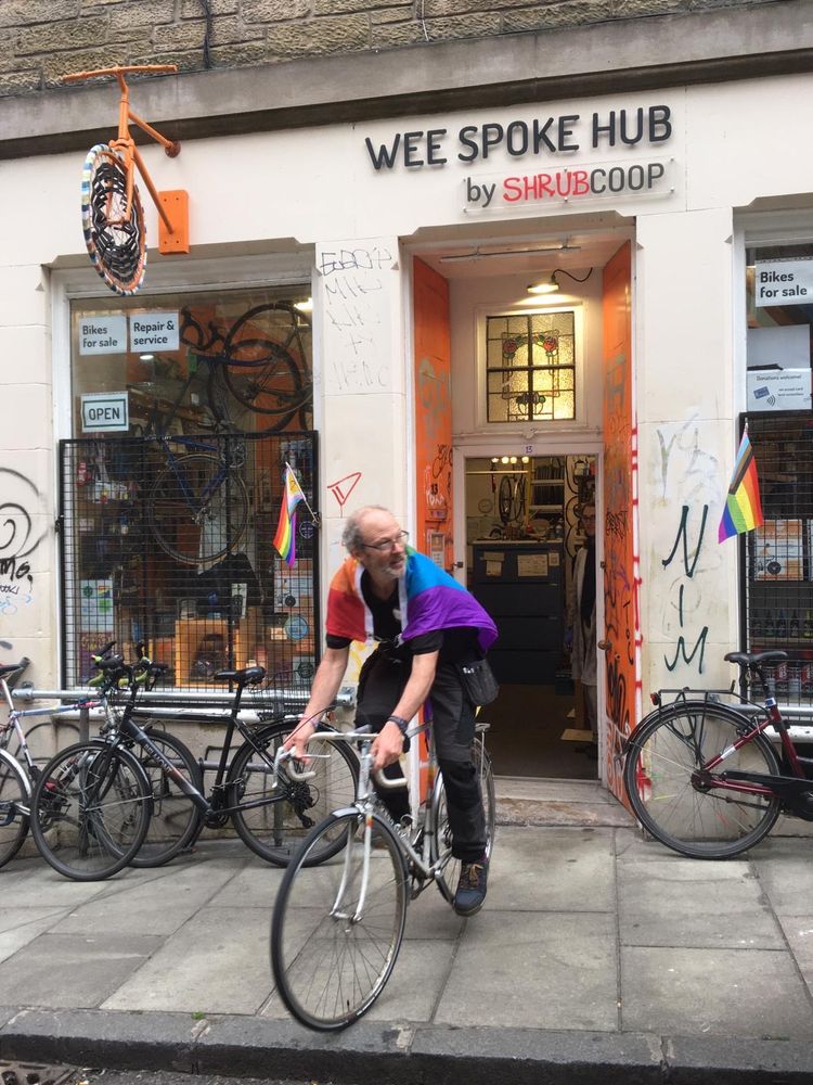 A cycle mechanic emerges from the door of the Wee Spoke Hub, Edinburgh's inclusive cycle workshop, test-riding a white Peugeot drop-bar bicycle. He is draped with a Pride flag, in the style of a Superman cape.  More Pride flags are visible in the background - it could be that the photo is taken on Pride Weekend.