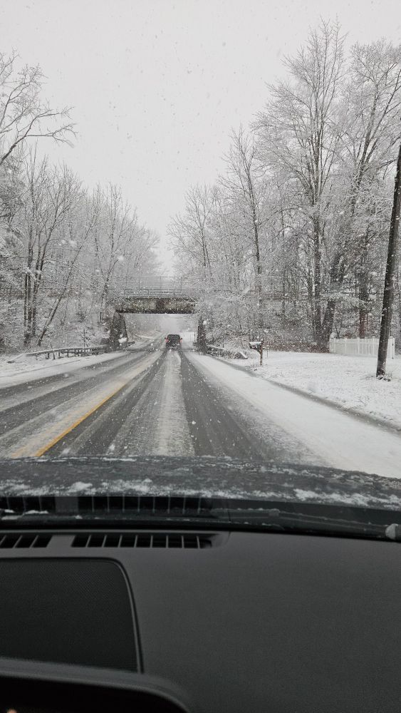 A snow-covered road with a pickup truck and overpass in the distance. Grass and trees are also covered. Some large snowflakes are visible. 