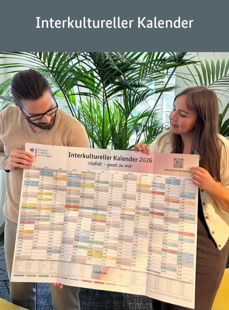 Zwei Personen halten gemeinsam einen großen Wandkalender mit der Aufschrift „Interkultureller Kalender 2026 – Vielfalt passt zu mir“. Oben ist das Logo des Bundesamts für Migration und Flüchtlinge zu sehen. Im Hintergrund stehen grüne Pflanzen.