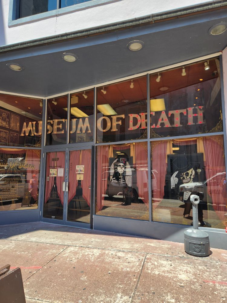 Glass storefront for Museum of Death. There are red curtains in the windows as well as artwork of skeletons in fancy dress.