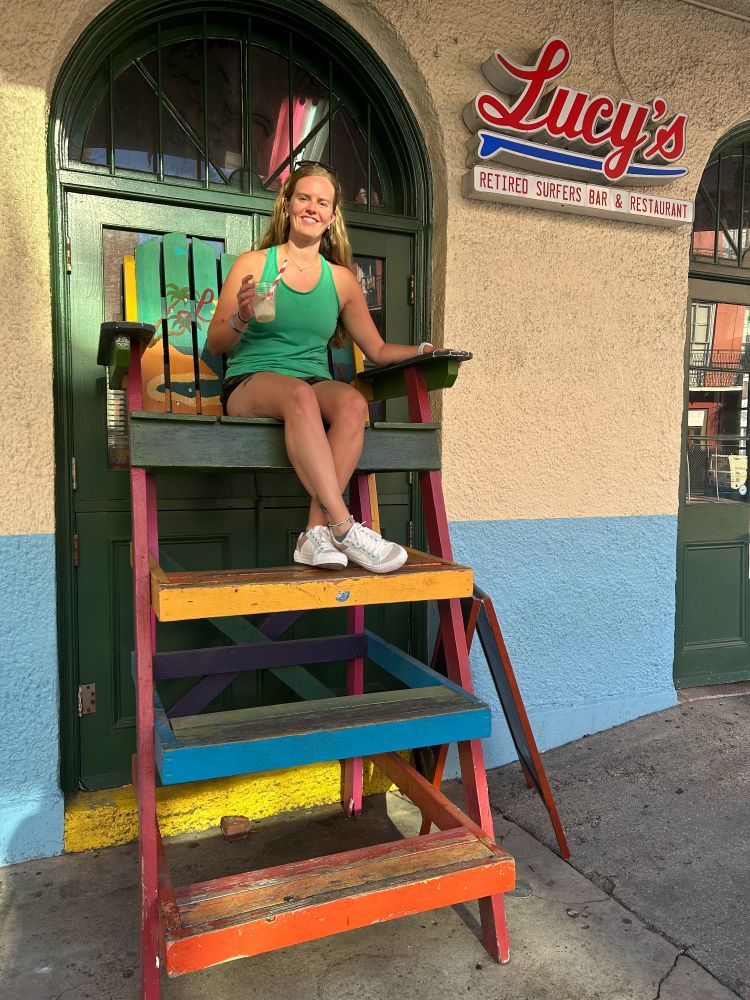 A woman sitting in a tall beach chair outside a restaurant named Lucy' Retired Surfers Bar & Restaurant. 