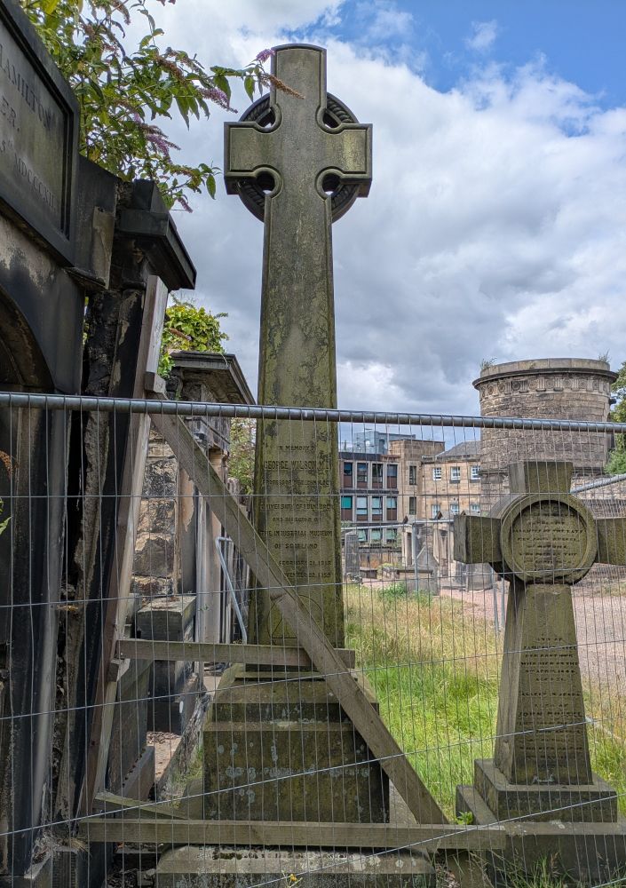 Memorial in the shape of a Celtic cross in a graveyard. it has some fencing and wooden supports in front, which are holding up the wall beside it.