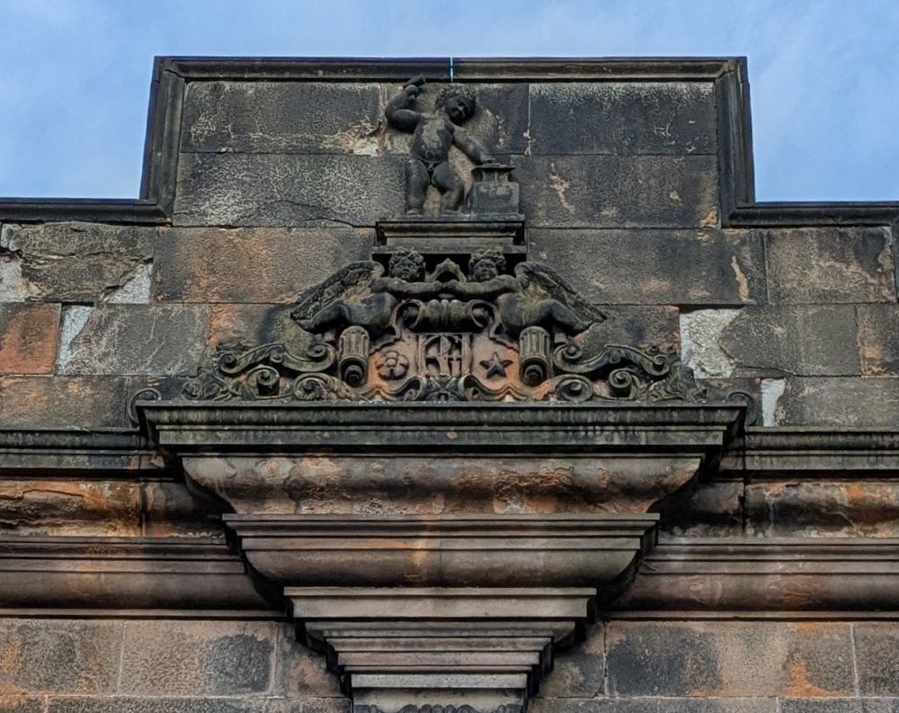 Detail cropped from the previous photo, showing a stone carving of putti welding a hammer over an anvil. Below are two winged cherubs, monogram and decorative stonework