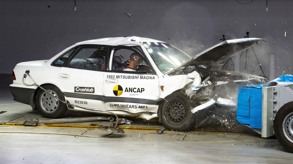 There's a white crash test dummy in the front passenger seat of a very crashed white 1993 Mitsubishi Magna. On the front door panel of the car is the black and yellow logo of "Ancap."