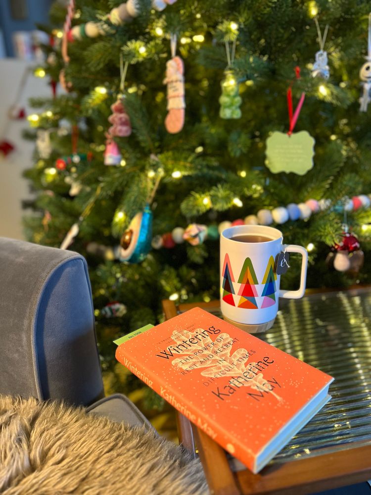 Image of a book, a cup of tea and a decorated lit up Christmas tree. Book title is Wintering by Katherine May 