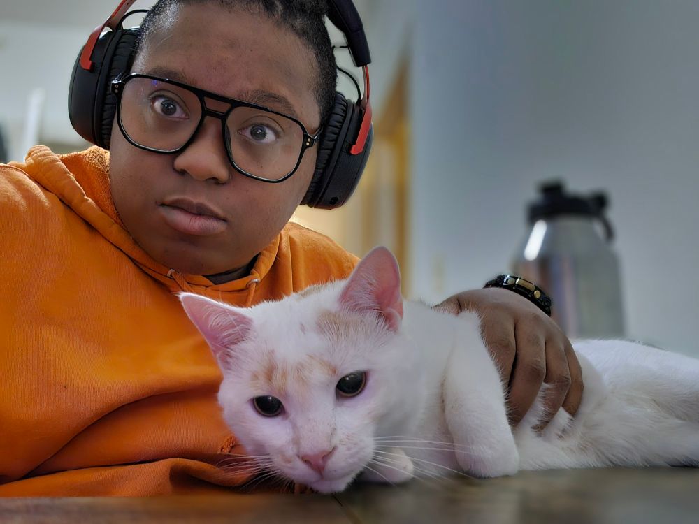 A human with medium brown skin wearing a bright orange hoodie and wearing glasses and headphones. They are holding a mostly white cat on a brown desk. There is a large water bottle in the background.