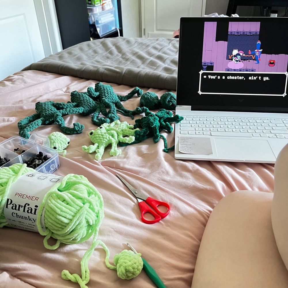 A bunch of various green crocheted frogs on a pink comforter. There’s a laptop playing a stream of the video game Deltarune next to the pile of frogs. There’s also a pair of scissors and a frog being made in the progress next to human knees.
