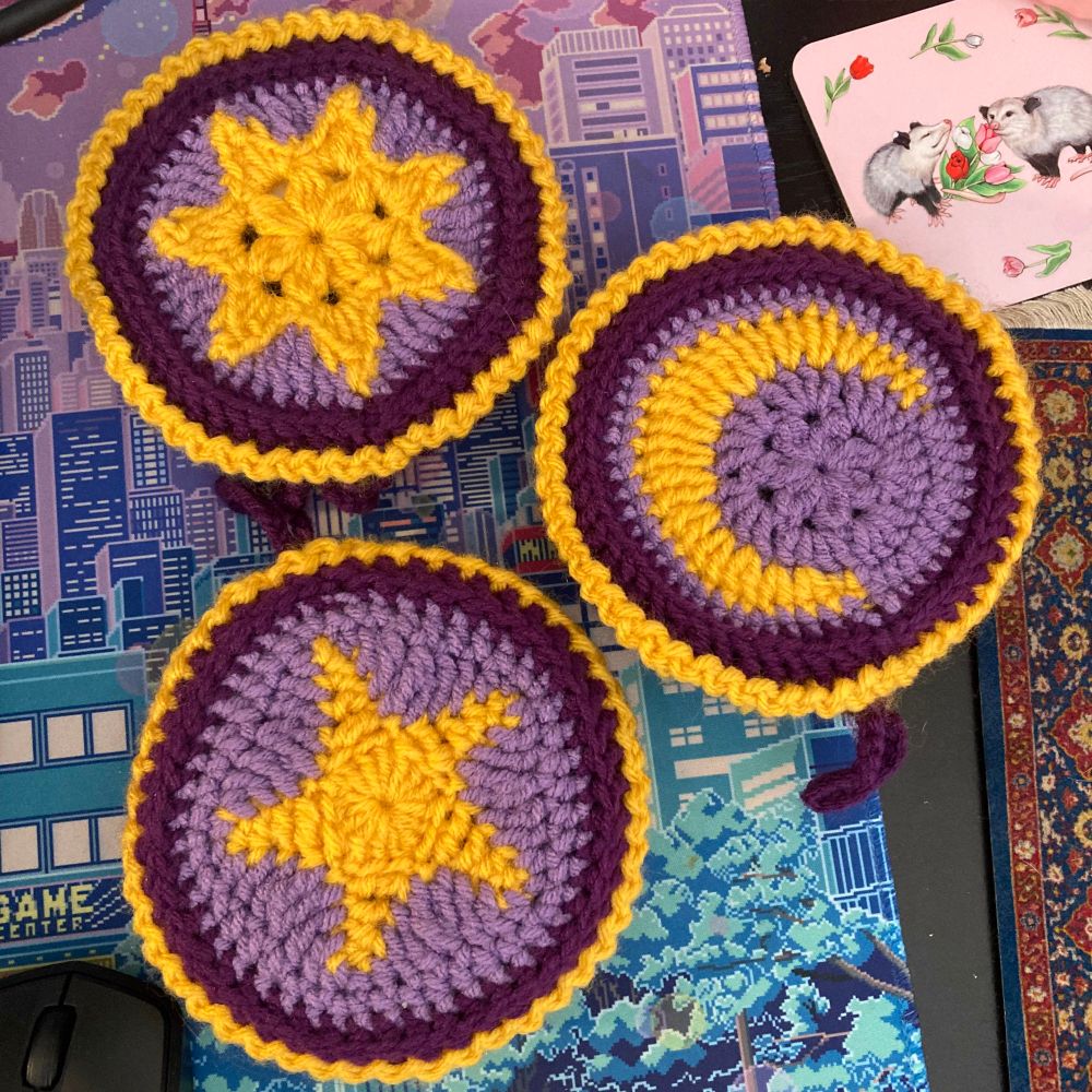 Three crocheted pill box holders on a desk. All are various shades of purple and yellow decorative borders with one having a yellow sun motif, one having a yellow moon motif and the third having a yellow star motif.
