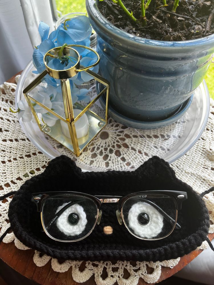 A crochet glasses holder that resembles the Sanrio character Chococat sits on a small side table with a couple of potted plants. There’s a pair of brow line glasses in the glasses holder.