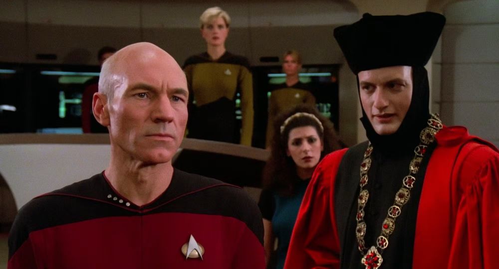 A still from Encounter at Farpoint, the first episode of Star Trek: The Next Generation, in which Captain Picard is standing on the Enterprise bridge looking very seriously at something serious offscreen while Q wears a funny hat and taunts him.  Officers Deanna Troi and Tasha Yar and an expendable crew member look on with concern.