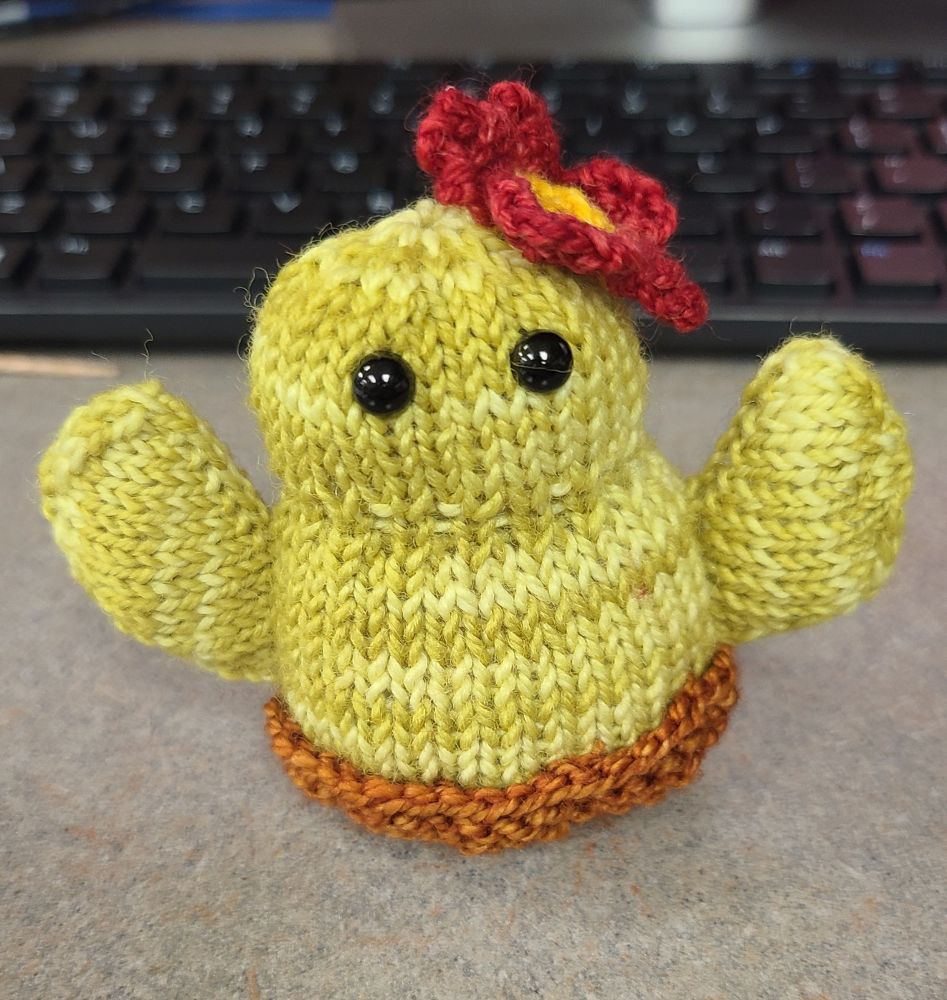 Emotional support cacti, knit in light green yarn, in a small brown knitted planter dish, with a red and yellow flower on the top of the cacti's head. 2 beady black safety eyes. 