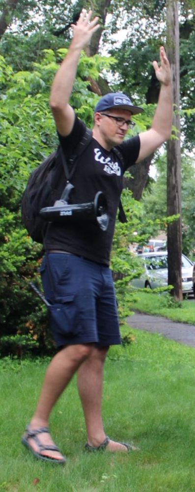 A tall man wearing sandals, navy blue shorts, a black “Wedge Live” T-shirt, and a blue Bryant Avenue hat holding his hands above his head