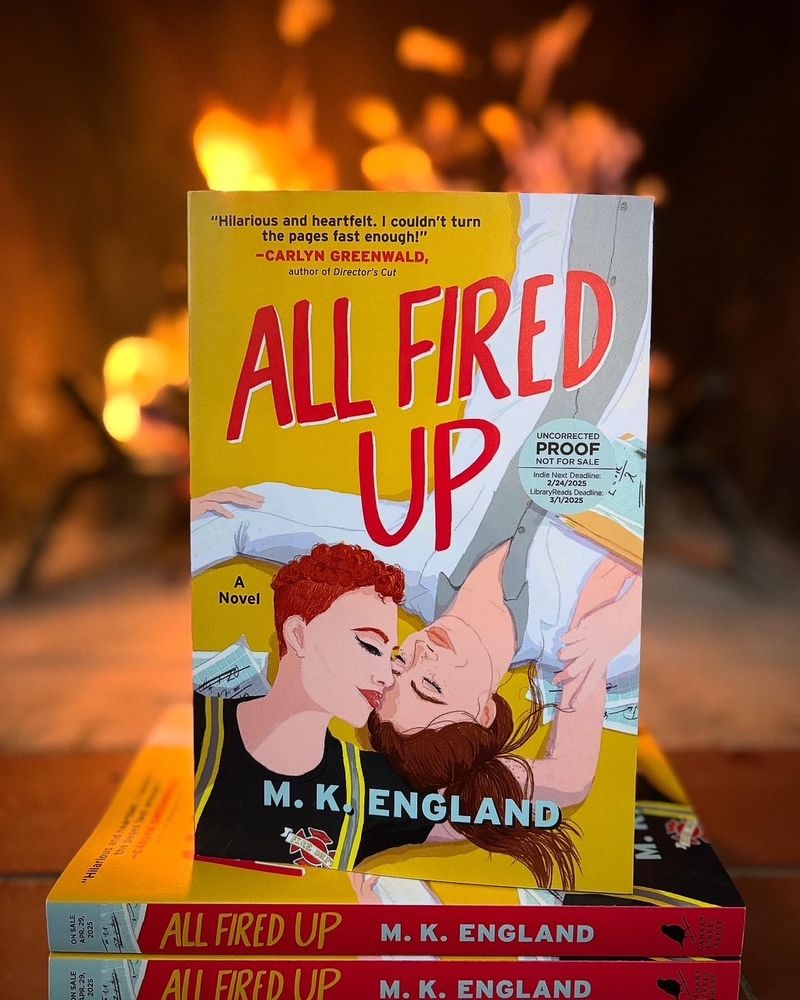 In the background is a lit fireplace with orange flames crackling. In front, two books lay flat, the spines reading ALL FIRED UP - M. K. England. One copy of the book stands face out on top of the stack, showing the yellow cover with red lettering. On it, two women lay in opposite directions, one nuzzling the other's forehead. One is wearing a firefighter uniform undershirt with bunker gear straps and the other wears a button down shirt with a white lab coat over it.