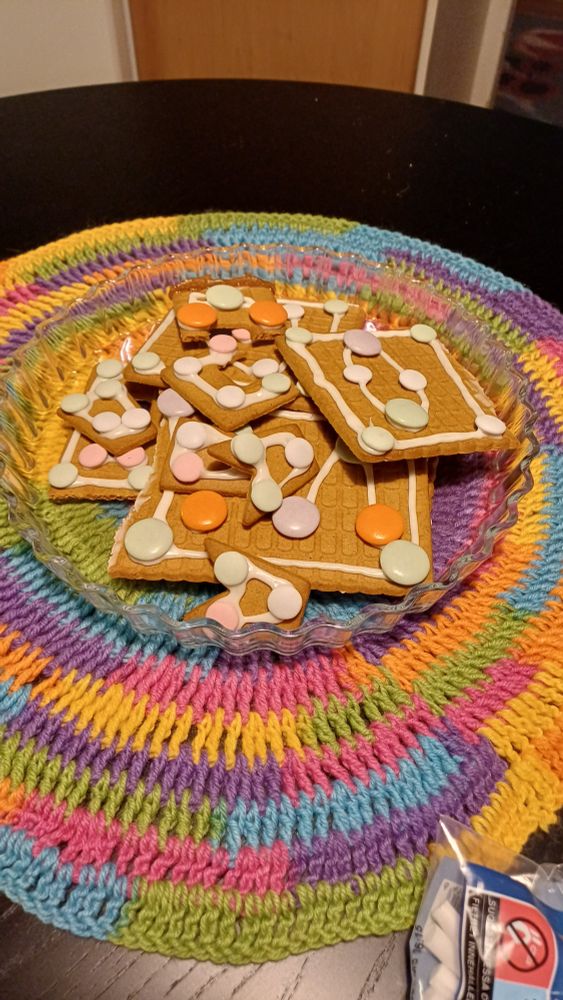 Kasa koristeltuja piparitalon osia sateenkaaren värisen virkatun pöytäliinan päällä. 