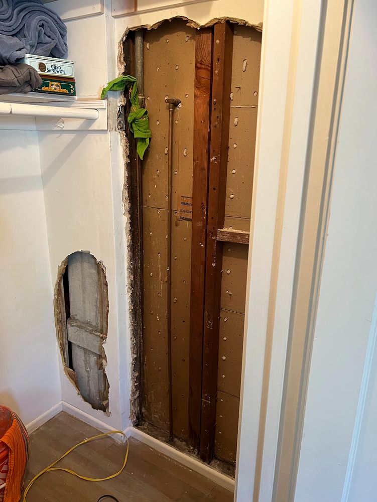 The inside of a deep square closet, with the plaster wall ripped out to expose two copper pipes and some studs. 
