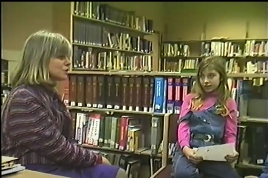 A screen cap from an old VHS tape - setting is in a library with a bunch of bookshelves. On the left is a white woman with grey hair cut to her shoulders; on the right I am about 9 years old, with long blonde hair, overalls, and a bright pink turtleneck.