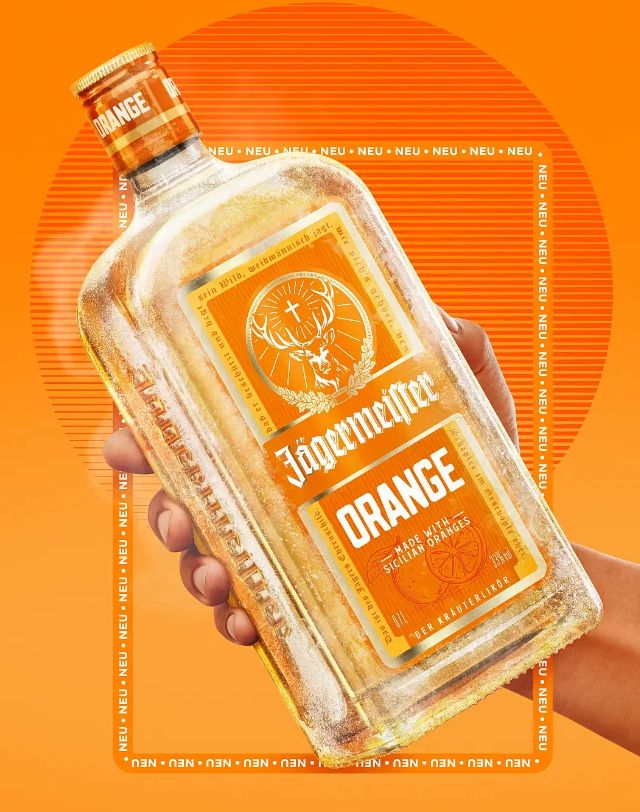 a person's hand holding a bottle of Jägermeister ORANGE, a new alcoholic beverage "made with sun-ripened Sicilian oranges and a light herbal note , 100% natural ingredients and masterful craftsmanship ensuring a balanced taste experience."