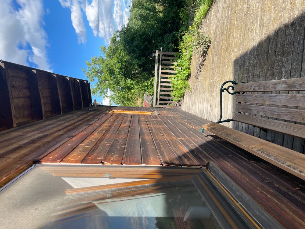 View along length of wooden extension: horizontal planks. Windows showing. With decking running along its length. A wooden bench sits next to the house facing the garden. Trees in distance. Garden to the right. Unseen, behind this view are the steps up to the seating area. 