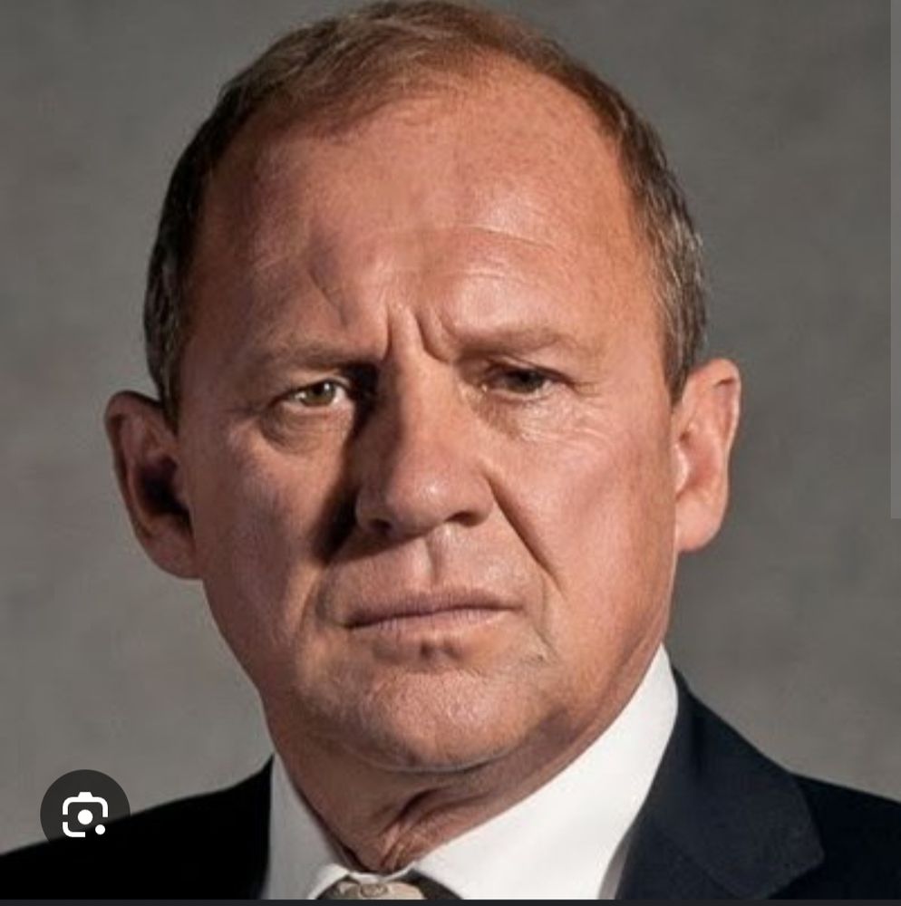 This is a still of the actor Peter Firth who played Sir Harry Pierce in the TV series Spooks. As a, spy, he has a serious and concerned expression and is wearing a black suit with a white shirt collar and a beige tie. 