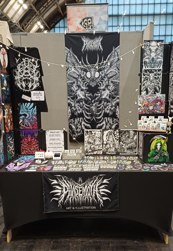 A photo of my table and display at MegaCon Manchester this weekend. A large moth banner is hanging up on the wall behind the table, and you can see prints, tees, stickers and bookmarks on the table. 