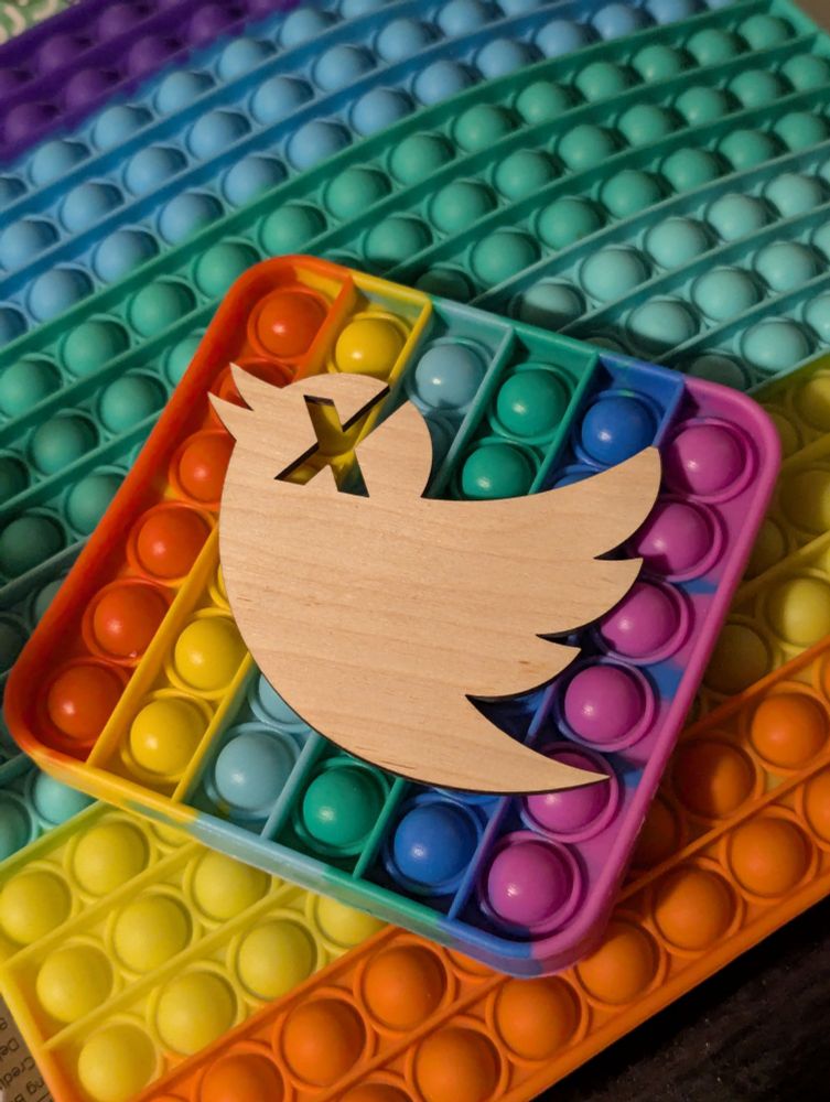 Laser cut thin wooden Twitter bird logo with an X through its head. The background is rainbow popper fidgets.