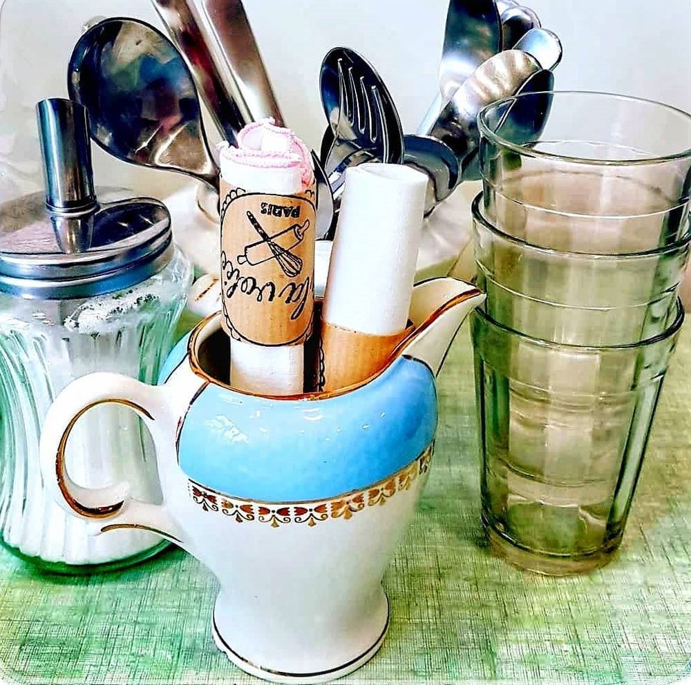 Sur une toile cirée verte, quatre verres de cantine empliés, en verre fumé, une sucrière en verre avec son distrubuteur en inox, un bouquet de couverts dans une tasse blanche en porcelaine. Et au premier plan, deux serviettes blanches enroulées et posées dans un pot à lait en porcelaine blanc et bleu ciel avec des détails dorés