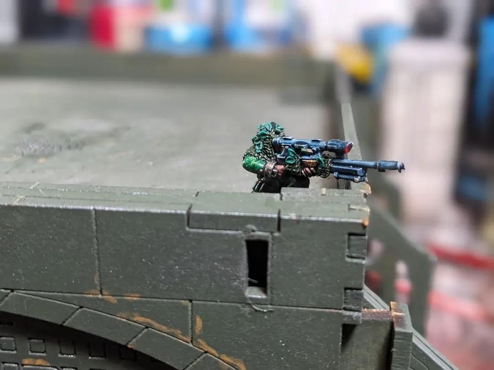 Photo. Painted Warhammer 40,000 miniature on a miniature building's flat rooftop. He balances his gun on a low wall. Model is photographed from the side and so you get more of a sense of his placement being high up on a building. The character Scope, kneeling, looking down the scope of a large sniper rifle. He's painted in shades of green, wearing camo and ghillie with brown leather boots, a dirty silver wrist manacle and tanned skin only visible on the hand. The gun is black with a gold winged skull emblem and a red scope lens.