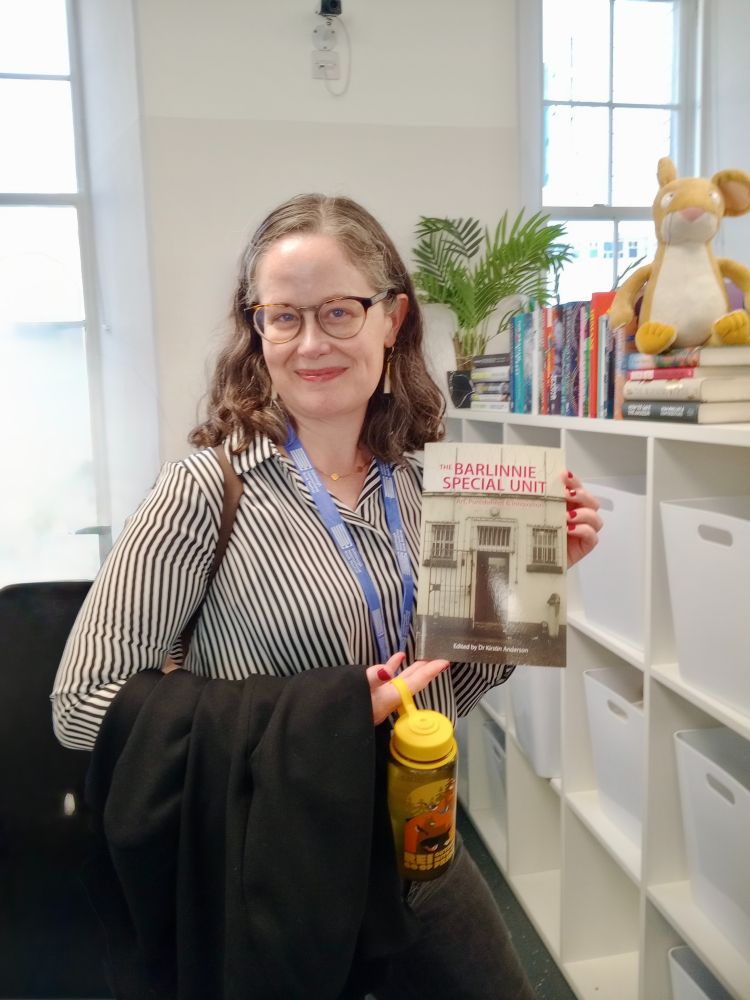 A woman wearing glasses and a striped black and white shirt smiling at the camera while holding a copy of the book "The Barlinnie Special Unit: Art, Punishment and Innovation"